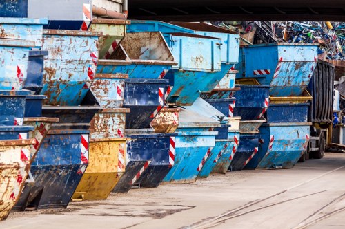 A row of large, industrial metal skip containers in varying shades of blue, with visible rust and weathering, lined up on a paved surface. The skips are partially filled with mixed waste materials, and some have open tops revealing debris inside. They are positioned along a curb or driveway area, with a shadow cast by overhead structures, indicative of an outdoor waste collection or disposal site in the vicinity of Bounds Green. The environment shows signs of frequent use, with tire marks and dirt on the ground nearby, and the skips are arranged in an orderly manner as part of a commercial rubbish removal operation by Garden Clearance Bounds Green, serving local domestic or business rubbish clearance needs in the area.