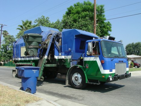 A large blue and green waste collection truck with a silver front, parked on a suburban street during daylight hours. The truck is equipped with an automated lifting mechanism at the rear, which is handling a green wheelie bin that is partially filled with waste, positioned on the curbside. The truck's side panel is open, revealing the interior hopper for waste collection. In the background, there are trees with lush green foliage, a utility pole with power lines, and a clear blue sky indicating fair weather. The truck's design and markings suggest it belongs to a rubbish removal service, likely for garden or general waste clearance, servicing local areas such as Bounds Green or nearby towns. The nearby pavement and residential surroundings reflect a typical UK urban or suburban environment, aligning with local rubbish collection services like those provided by Garden Clearance Bounds Green.