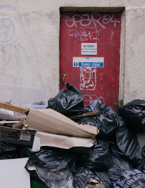 A pile of black rubbish bags and discarded waste materials, including cardboard and plastic packaging, stacked against a weathered red door set into a beige exterior wall. The door features graffiti t