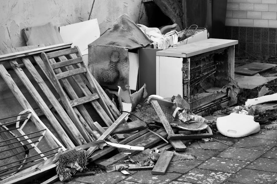 A pile of discarded household waste and debris located on a paved outdoor area next to a building wall. The pile includes a broken wooden pallet leaning against a large, bent metal object that appears