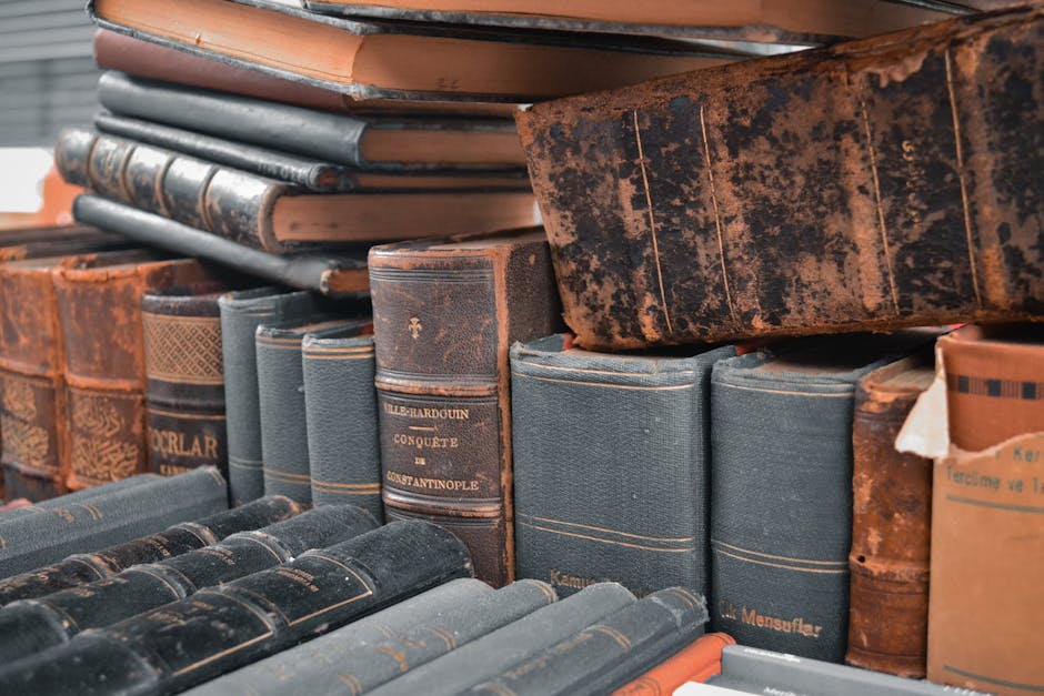 A close-up view of a collection of old, worn hardcover books stacked together, primarily featuring leather-bound volumes with varying shades of brown, maroon, and black. Some books display gold or emb