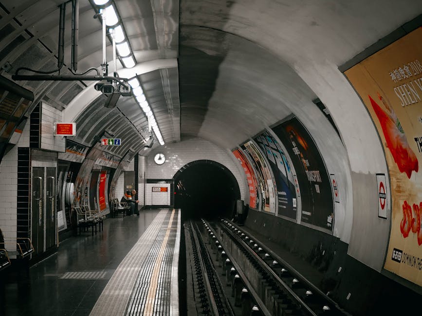 A close-up image of the London Underground roundel sign mounted on a light-colored brick wall. The sign consists of a circular red ring with a thin white border, enclosing a horizontal blue bar with t